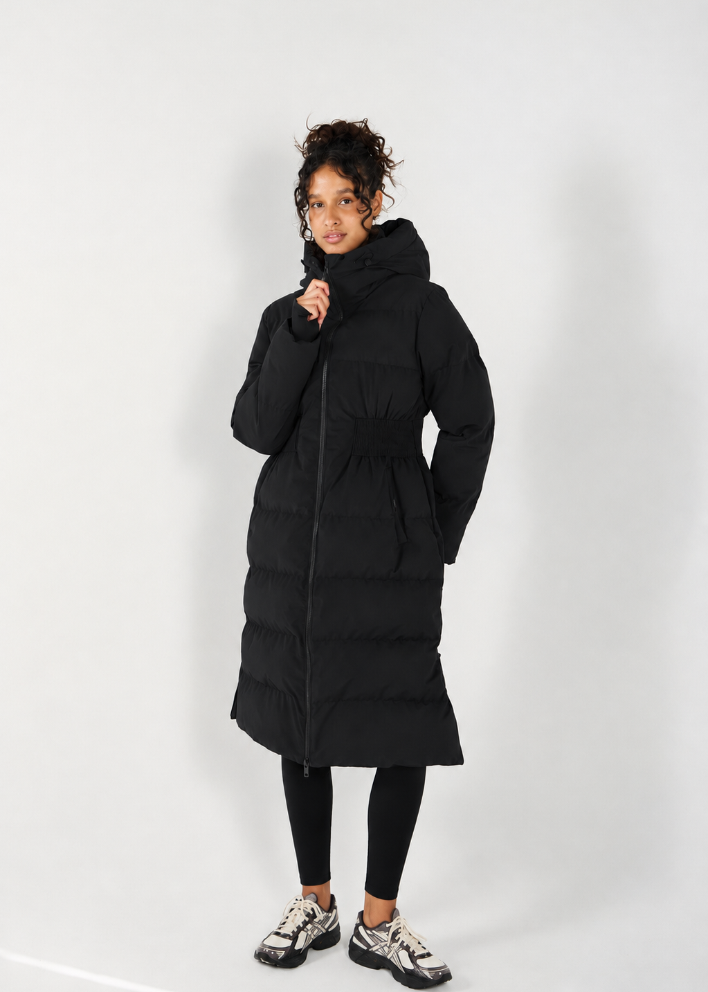 A model wearing a long black hooded puffer coat with a front zipper, photographed against a plain light gray studio background.